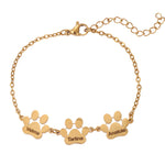 A gold-colored bracelet with engraved paw prints and the names 'Anna', 'Lena', and 'Tino' on a chain structure.